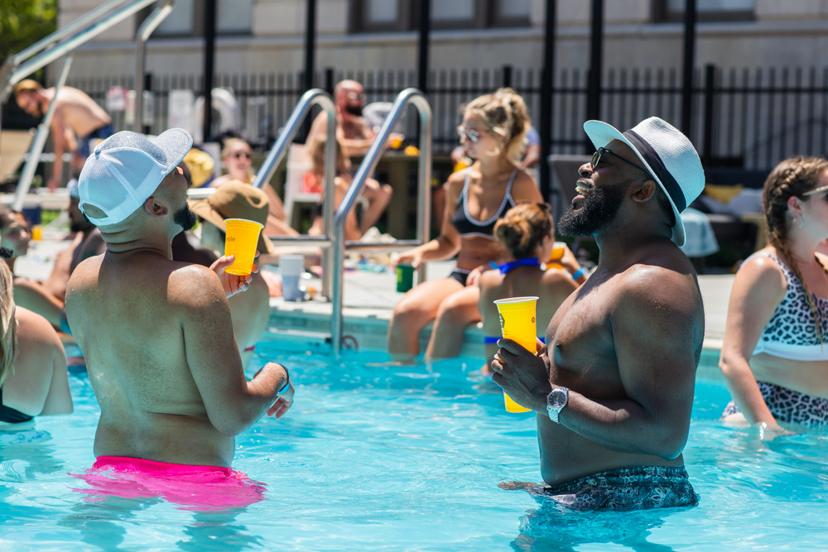 Two men in hats laughing and holding drinks in a busy outdoor pool, surrounded by people enjoying a sunny day.