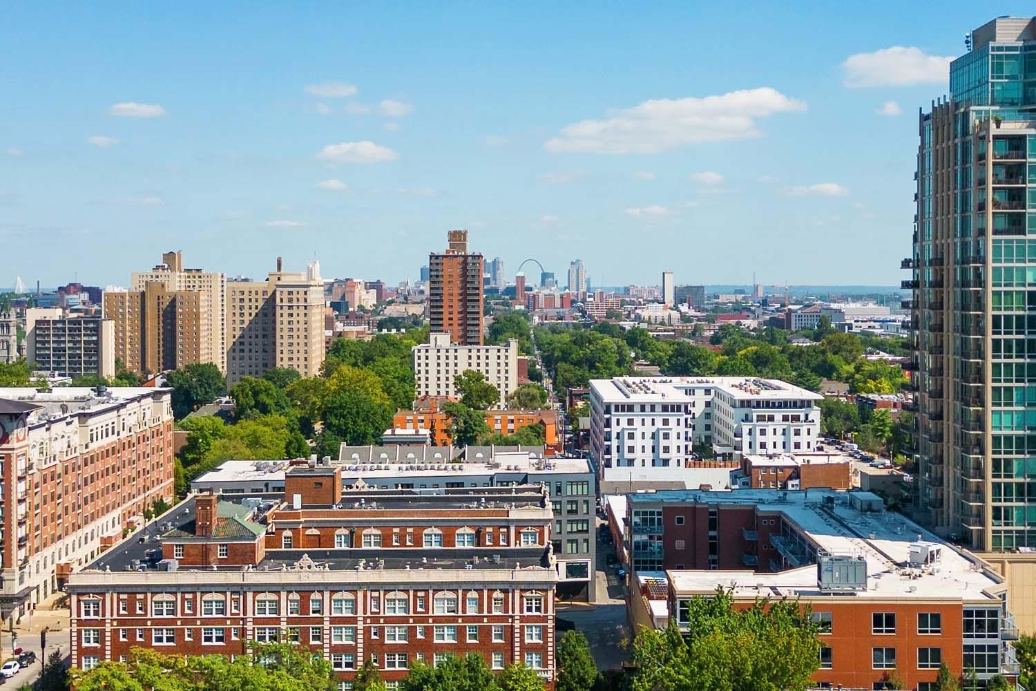 A cityscape view featuring buildings of varying heights, greenery, and a distant view of a skyline with a visible arch under a clear blue sky.