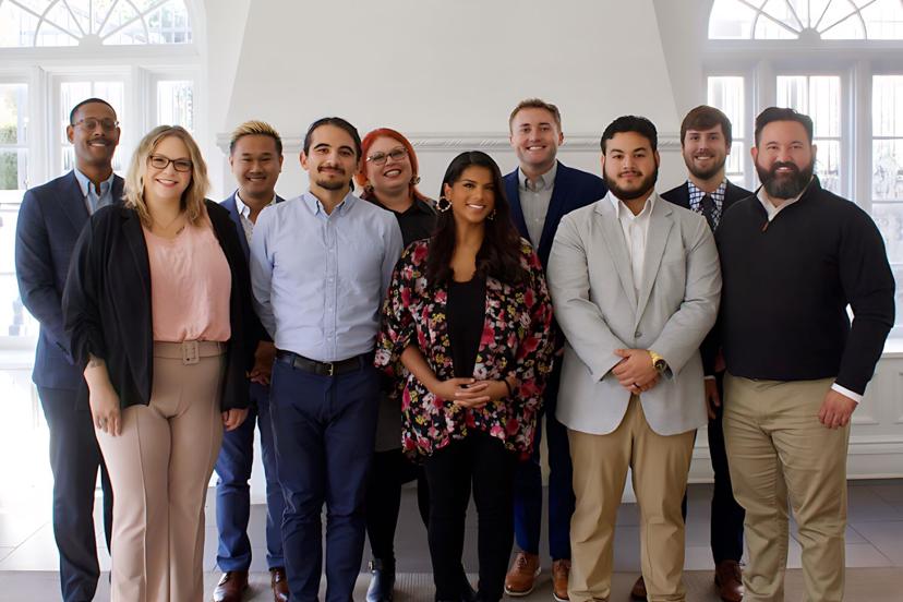 A diverse group of ten people, smiling, standing indoors in a well-lit room with white walls and large windows.