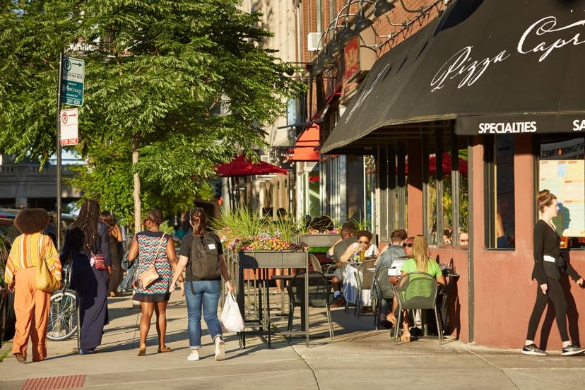 People walking on a sunny street next to a pizza restaurant with outdoor seating and plants.