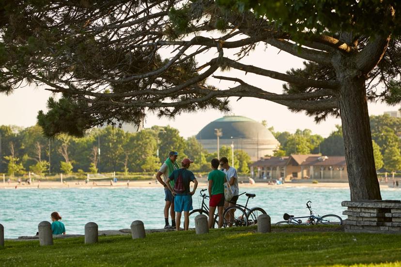 Group of people talking near bicycles beside a lake, with a beach, trees, and a dome-shaped building in the background.