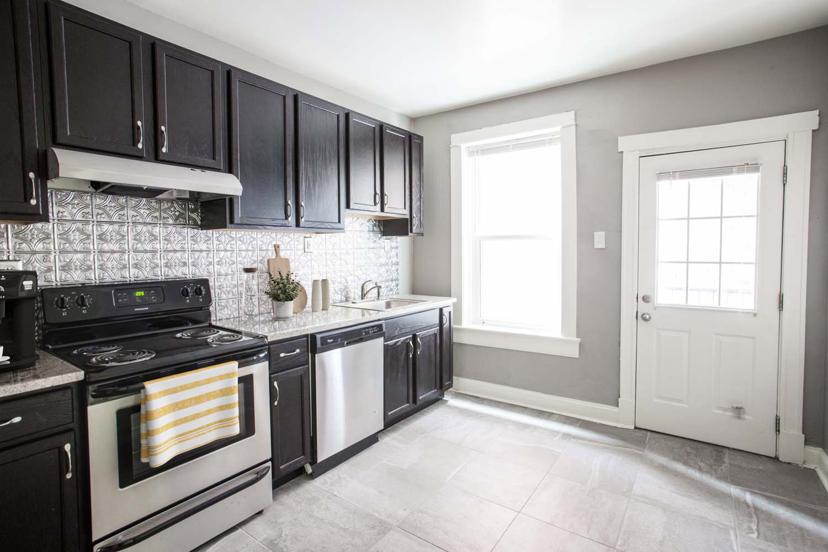 Modern kitchen with dark cabinets, stainless steel appliances, tiled backsplash, and a large window next to a white door.