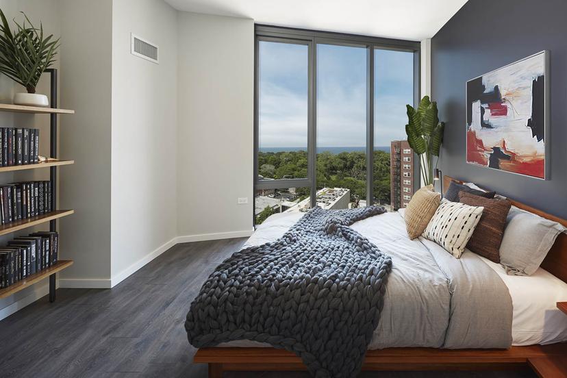 Modern bedroom with a large window, abstract painting, cozy bed with pillows and a knitted blanket, bookshelf, and scenic city and tree view.