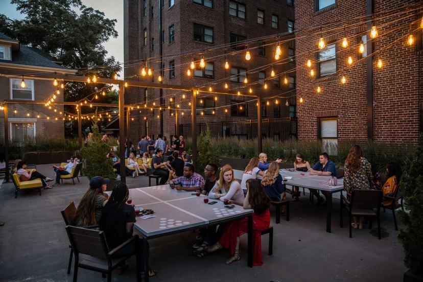 People gather under string lights at outdoor tables near a brick building, socializing during early evening.