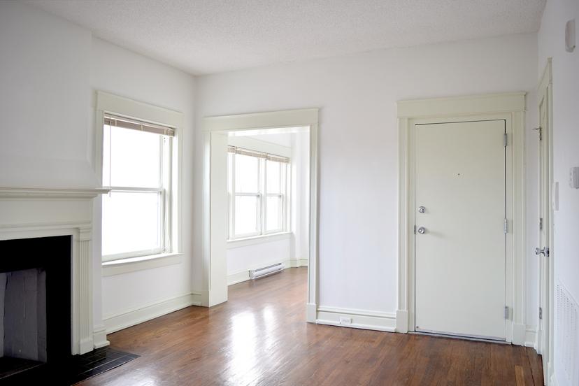 Bright, empty room with wood floors, white walls, a fireplace, and large windows. An entrance door is on the right.