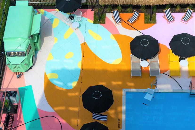 Aerial view of a colorful poolside area with a retro green van, black umbrellas, striped loungers, and vibrant abstract patterns on the deck.
