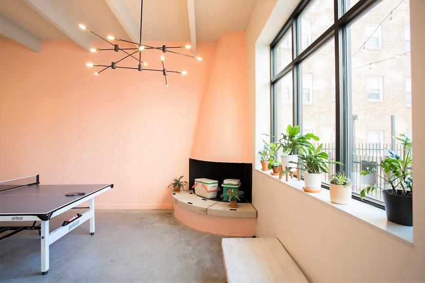 Bright room with peach walls, a round corner fireplace, potted plants on a window sill, and a ping pong table under a modern chandelier.