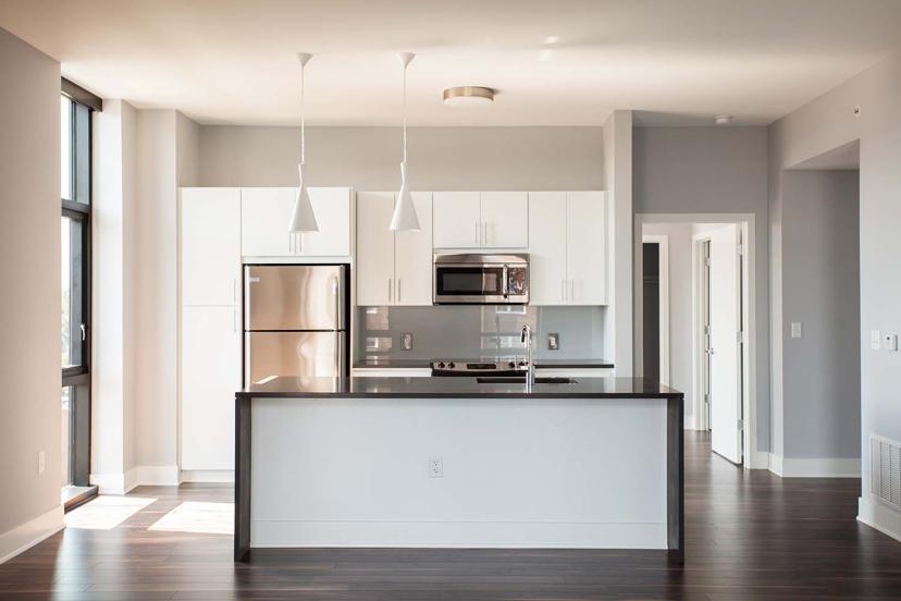 Modern kitchen with white cabinets, stainless steel appliances, dark countertops, pendant lights, and hardwood floors, illuminated by natural light.
