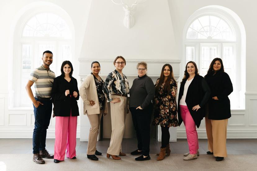 A group of eight people standing together indoors in front of a white wall with arched windows, smiling and posing for a photo.