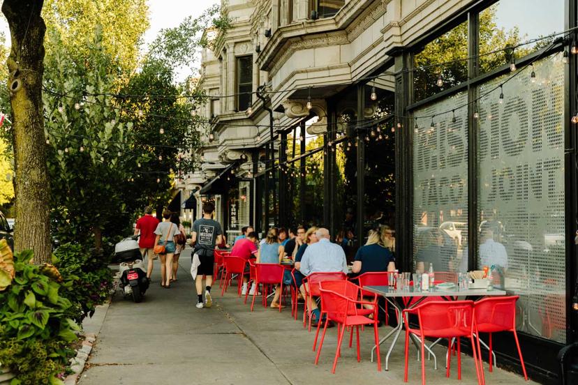 People dining at red tables outside a cafe on a tree-lined street, with string lights overhead and a decorative building facade.