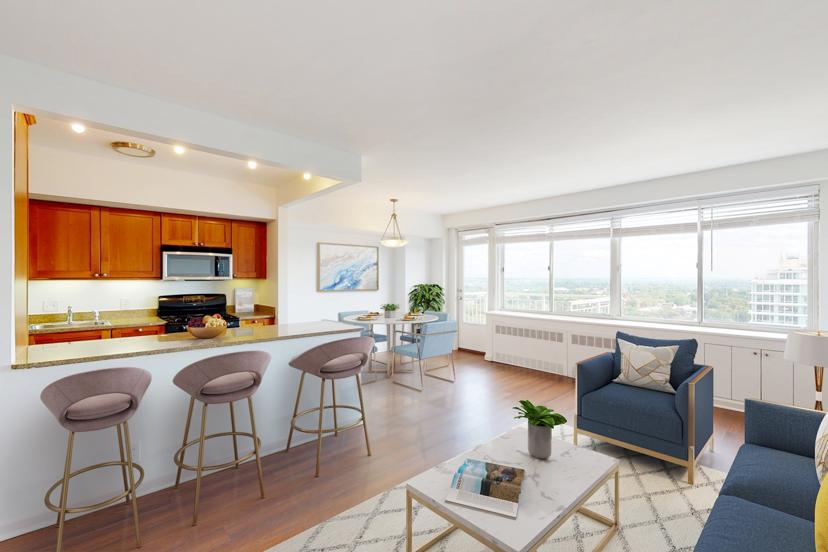 Modern open-plan living room with a kitchen, dining area, and large windows. Features include bar stools, a sofa, and a table with a plant.