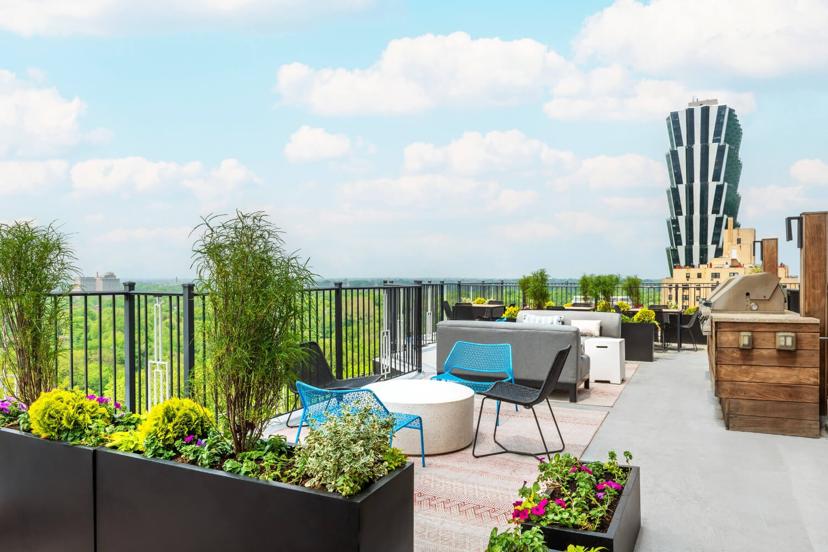 Rooftop terrace with colorful chairs, plants, and a barbecue grill, overlooking a scenic view with a unique building and lush greenery in the background.