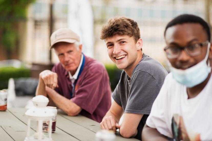 Three people sitting at an outdoor table, one smiling at the camera, another wearing a cap, and the third wearing glasses and a downturned face mask.