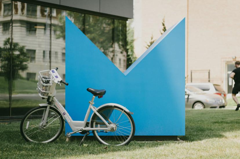 A white rental bike is parked on grass in front of a large blue sculpture shaped like an angular letter "M" beside a building with glass windows.
