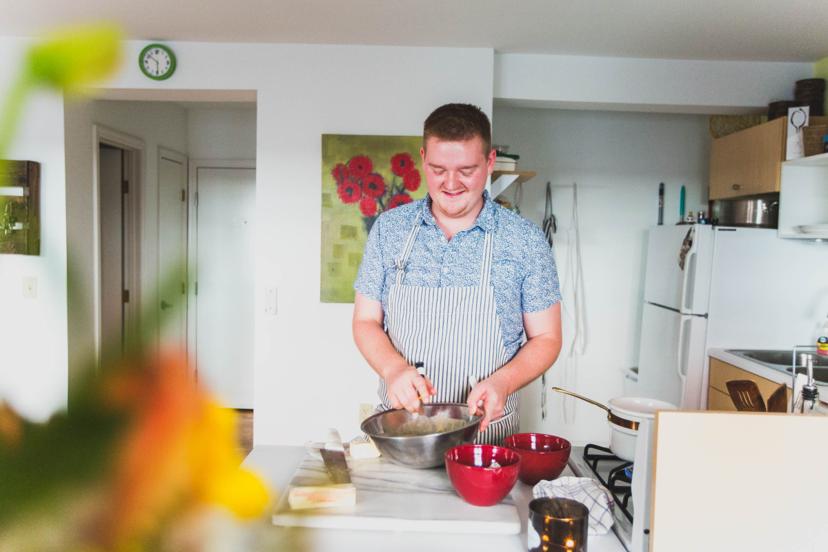 A person in a striped apron mixes ingredients in a bowl in a bright kitchen, surrounded by red bowls and cooking tools.