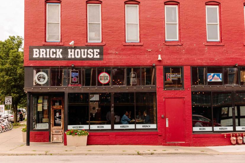 Red-brick building labeled "Brick House," with large windows, neon beer signs, a red door, and potted plants. People are seated inside.