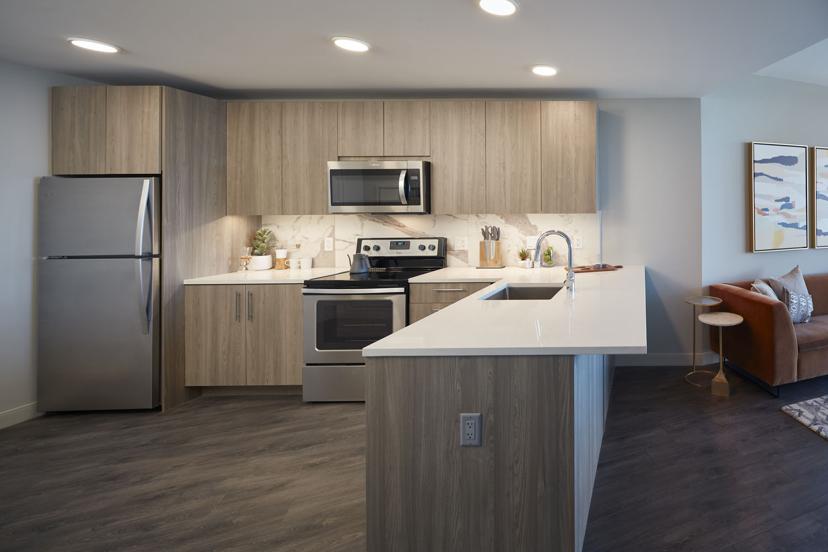 Modern kitchen with light wood cabinets, stainless steel appliances, and a white countertop. A cozy orange sofa and art prints are in the corner.