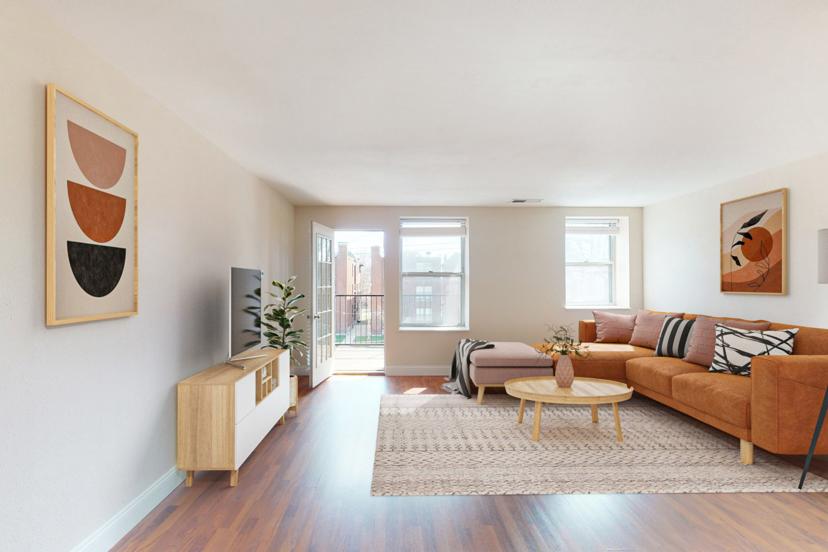 Modern living room with a tan sofa, patterned rug, coffee table, and wall art. Bright light from windows and a door to a balcony.