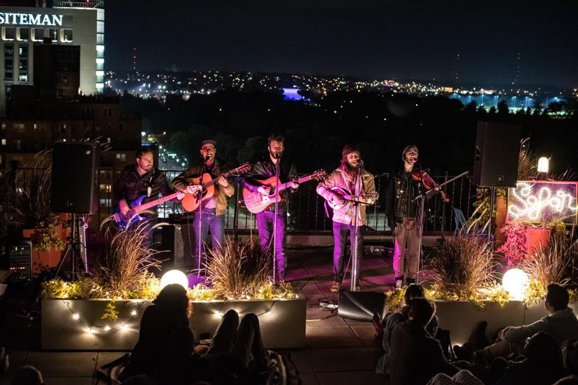 A band performs on a rooftop at night, with guitarists, a bassist, mandolin player, and violinist. City lights twinkle in the background.