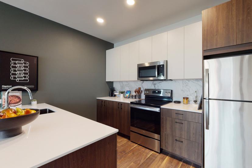 Modern kitchen with white countertops, stainless steel appliances, dark wood cabinets, and a bowl of fruit on the island.